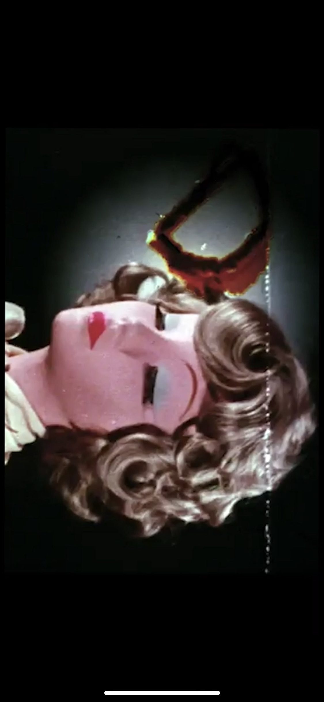 A film calibration frame known as a leader lady, turned on its side: a mannequin head with a pale pink face, closed eyes, bright red painted lips and a curled brown wig lies against a dark background. Above the head, a burn hole has eaten through the film, its edges scorched red and orange, and a line of tiny white specks runs down one side of the frame.